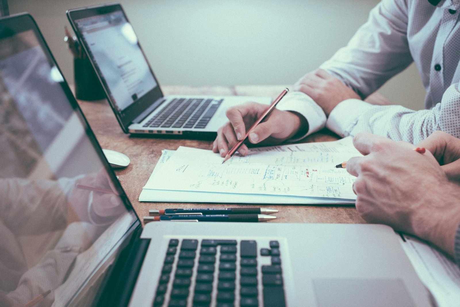 Two people at a desk with laptops, one writing notes on a printed plan with a pencil