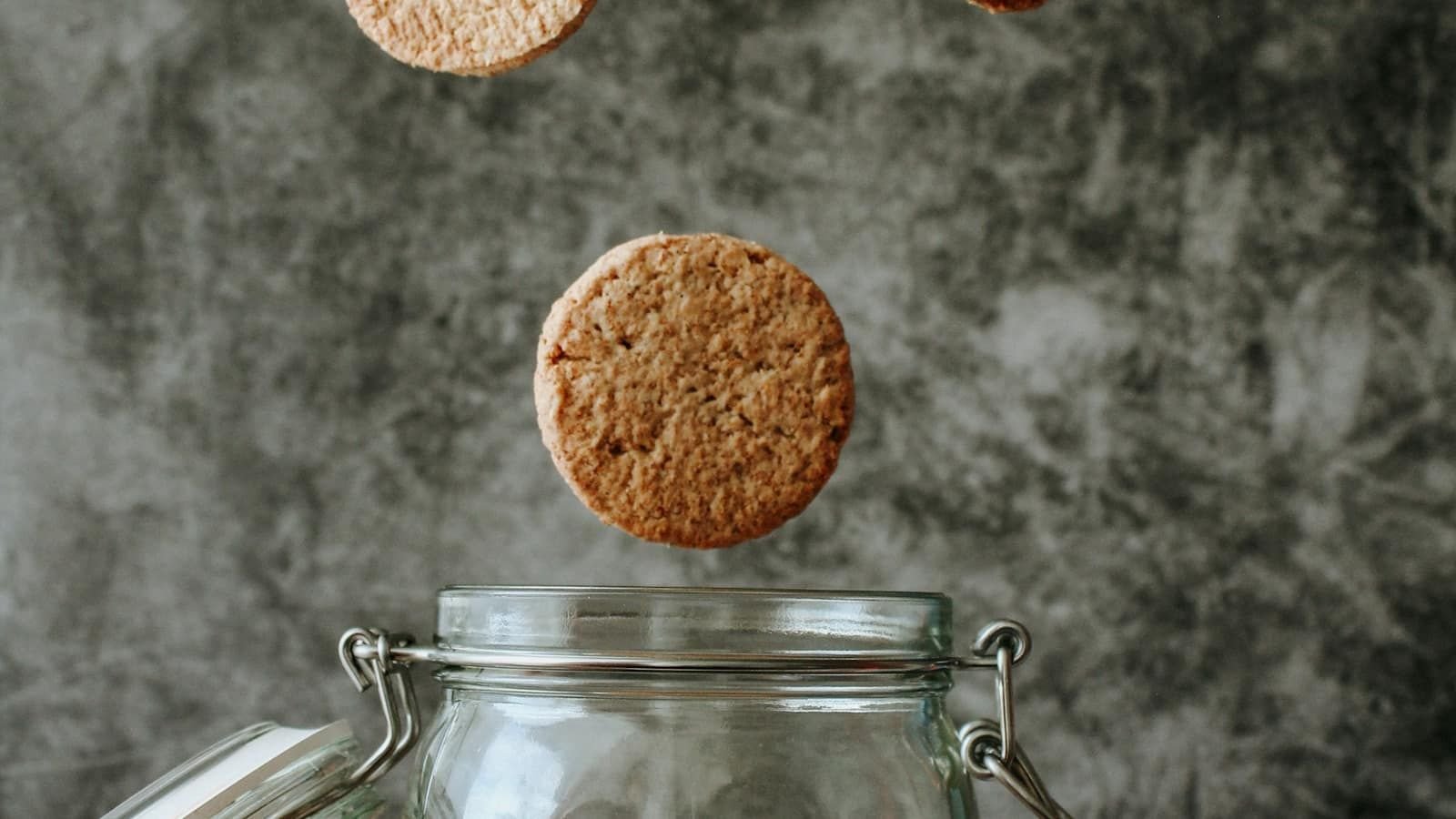 Round biscuits dropping into an open glass jar against a grey wall