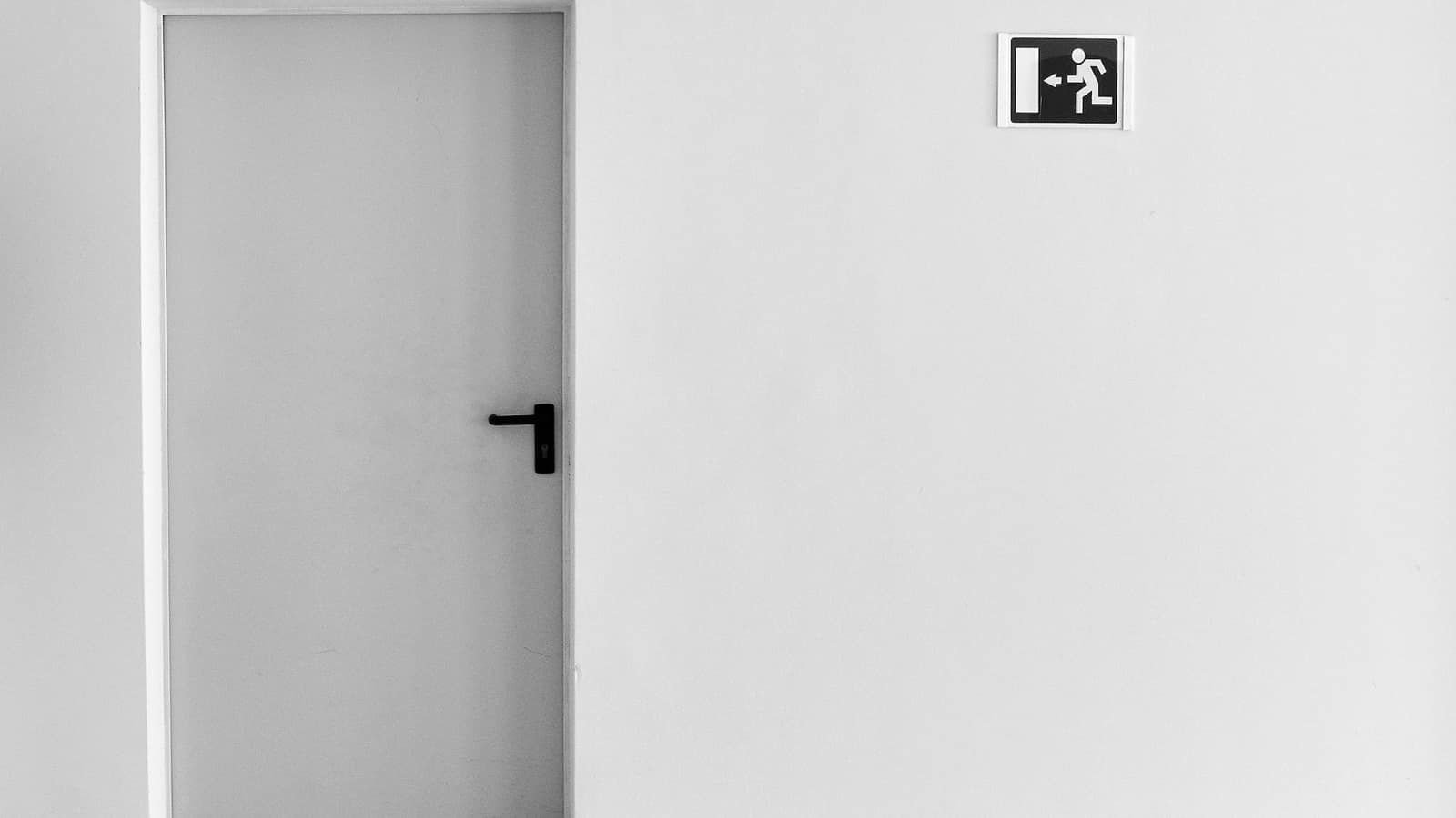 A plain grey door in a white wall with an exit sign above it