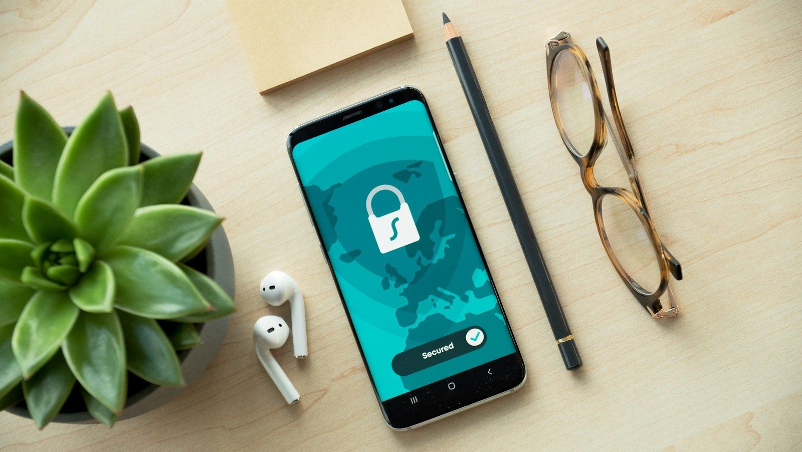 A smartphone showing a padlock icon on a wooden desk next to earbuds, a pencil and a plant