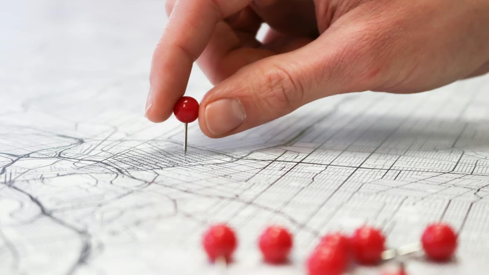 A hand pushing a red pin into a printed street map, with more red pins below
