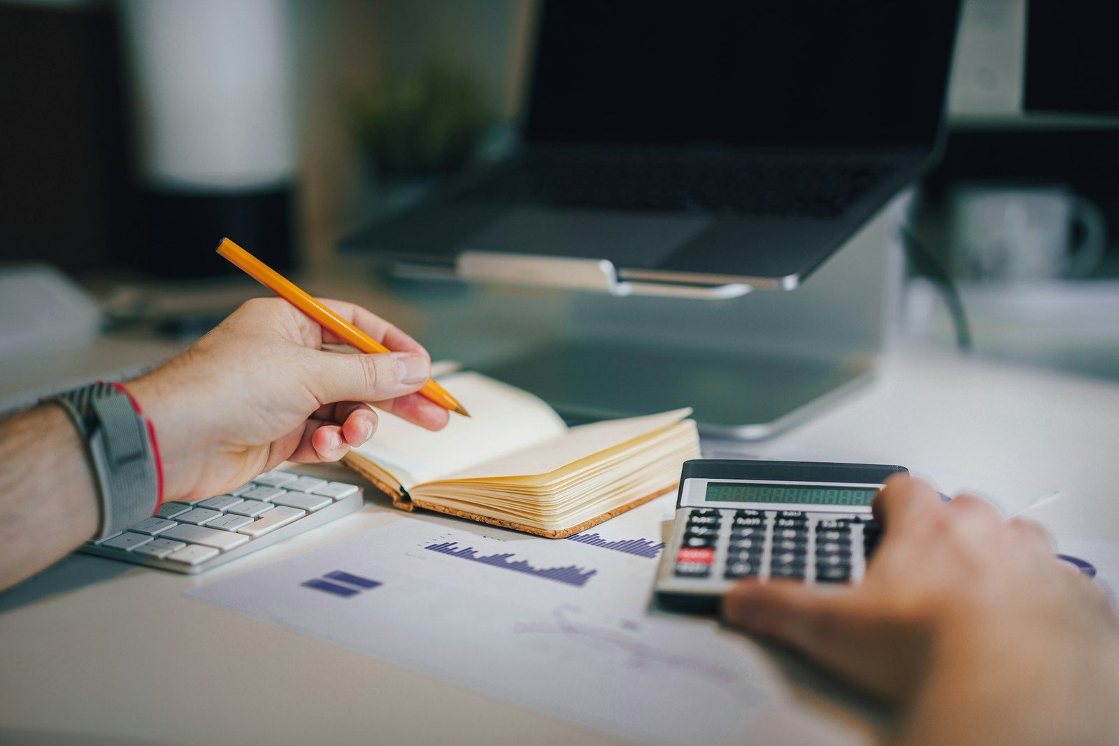Hands working at a desk with a calculator, an open notebook and printed charts beside a laptop