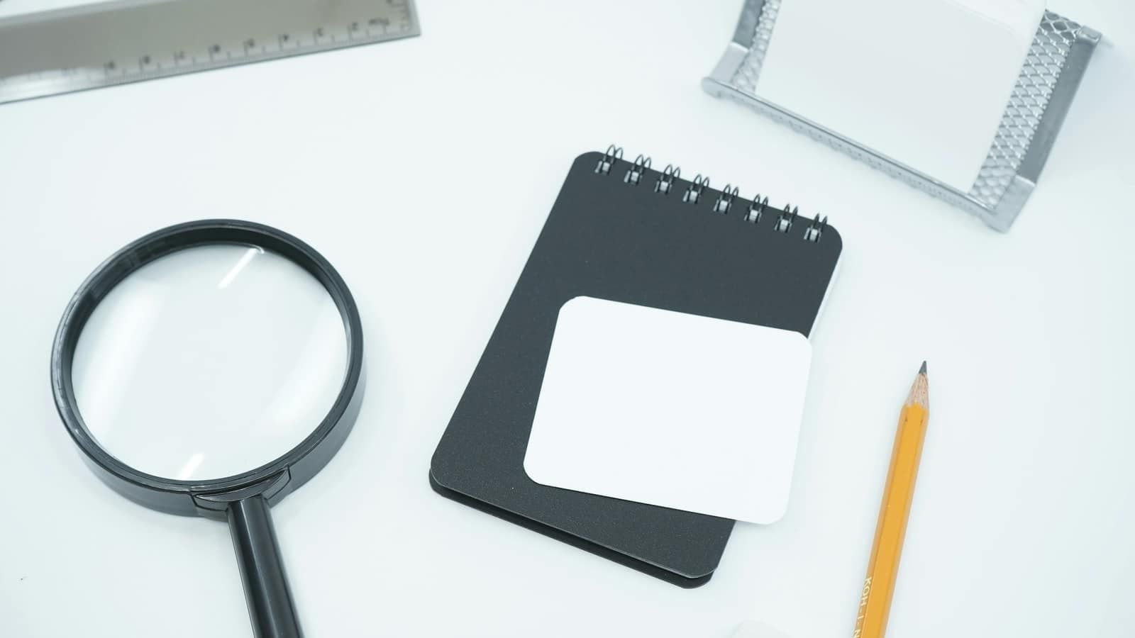 A black magnifying glass next to a notebook and pencil on a white desk