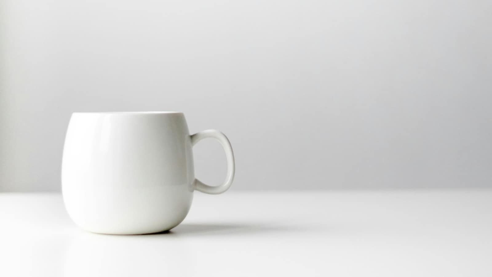 A plain white ceramic mug on a white table against a white wall